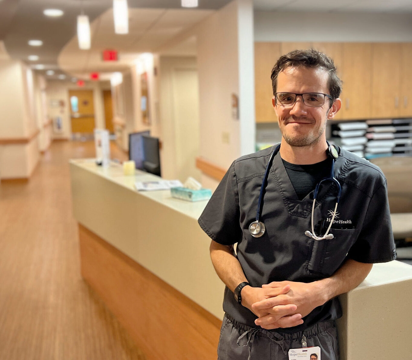 A male nurse stands against a nursing station in a warmly lit hospice center