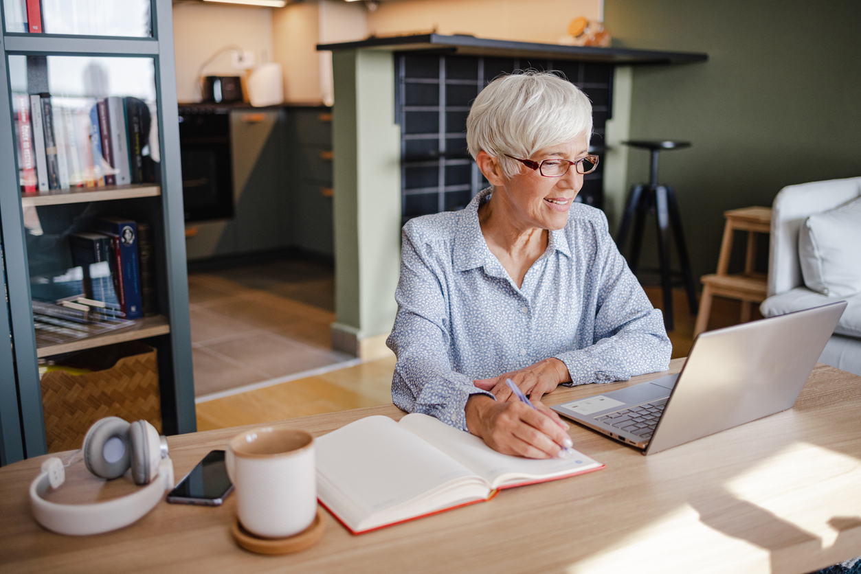 Senior woman is at home, she is using a laptop and writing notes in a notebook