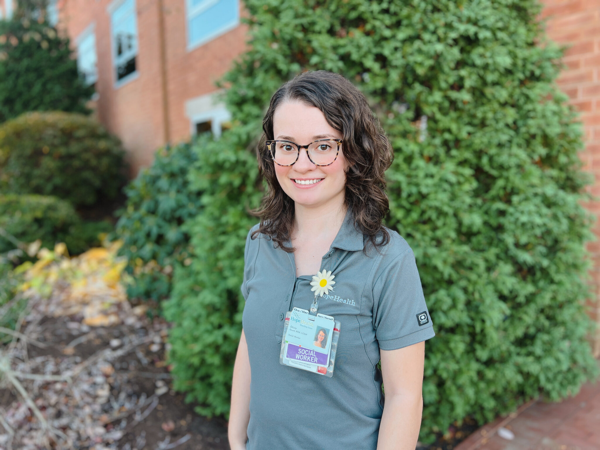 Smiling woman with curly brown hair wearing glasses and a gray HopeHealth polo shirt, standing outside in front of greenery. She is wearing a name badge and a daisy flower pin.