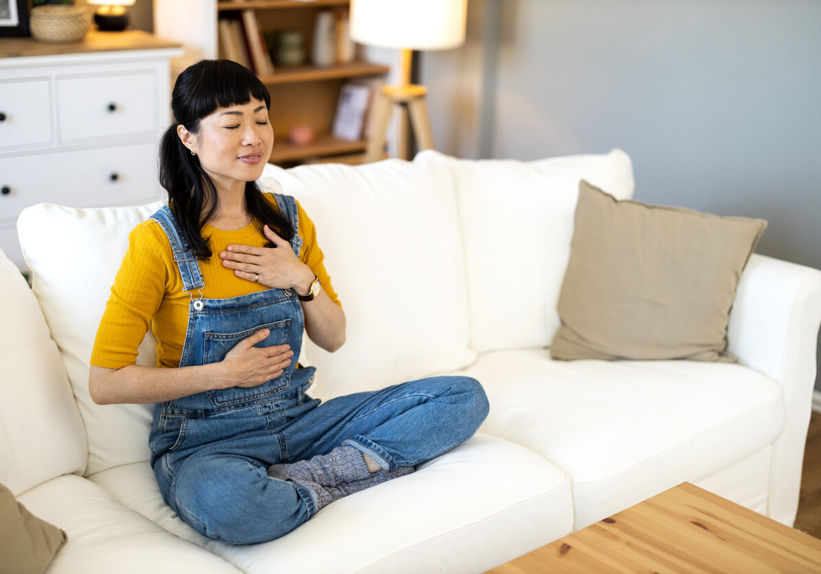 Shot of happy mid adult Asian woman practicing meditation in the living room at home.